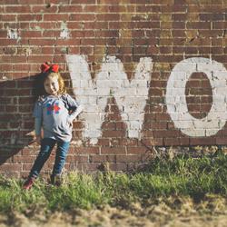girl standing in front of a red brick wall which has grafitti on saying "for women"
