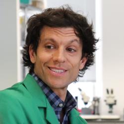 Dr Tim Weil in a green demonstrator's lab coat talking to year 12 students in the lab at an outreach event