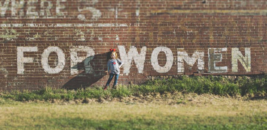 girl standing in front of a red brick wall which has grafitti on saying "for women"